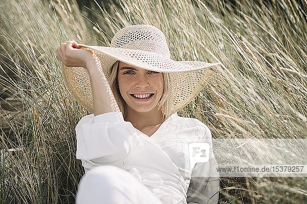 Porträt einer blonden jungen Frau in den Dünen mit weißer Wickelbluse und Sommerhut