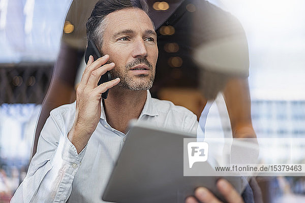 Porträt eines Geschäftsmannes mit Tablett am Telefon hinter einer Fensterscheibe in einem Cafe