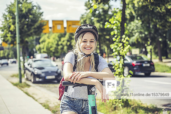 Porträt einer glücklichen jungen Frau mit Fahrradhelm  die sich an den Lenker eines E-Scooters lehnt