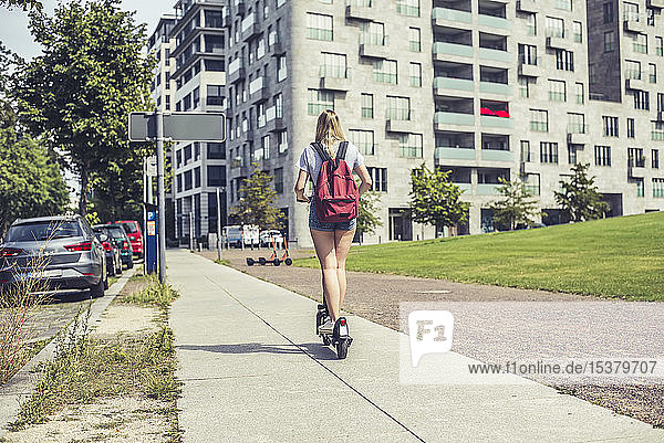 Rückenansicht einer jungen Frau mit Rucksack auf einem E-Scooter auf dem Bürgersteig  Berlin  Deutschland