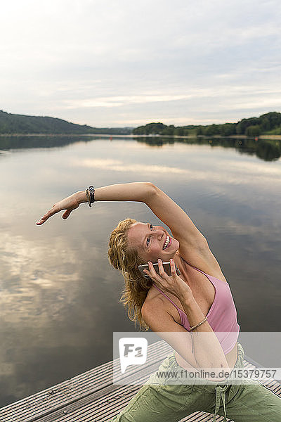 Junge Frau am Telefon beim Turnen auf einem Steg an einem See
