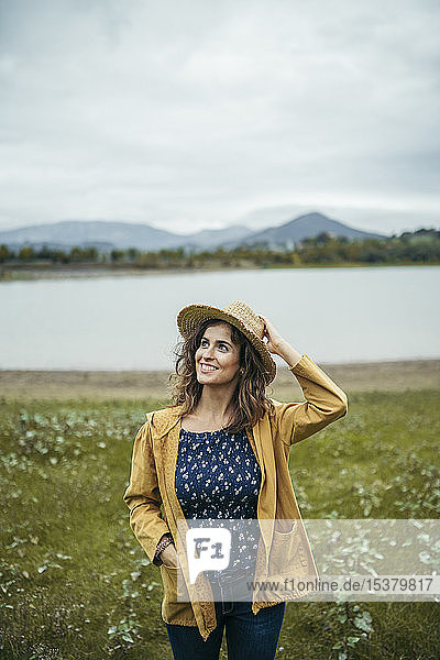 Happy young curly haired woman wearing a yellow coat and blue t-shirt holding a hat in her head with a lake in the background