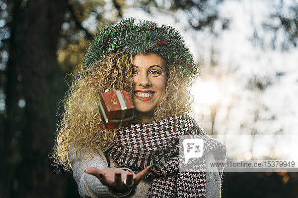 Porträt einer lächelnden jungen Frau mit Weihnachtskranz auf dem Kopf  die mit einem Weihnachtsgeschenk jongliert