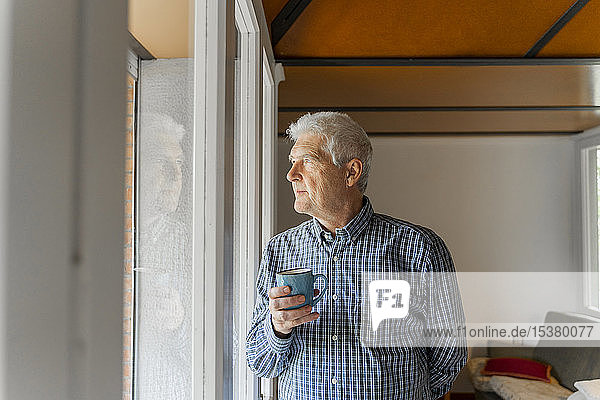 Ernsthafter älterer Mann  der durch ein Fenster auf sein Zuhause schaut