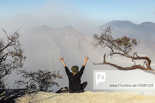 Frau mit Atemschutzmaske sitzt am Vulkan Ijen und hebt die Arme  Java  Indonesien