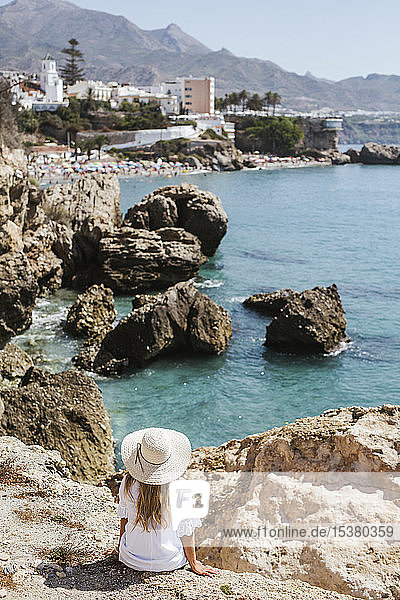 Rückenansicht einer Frau in weißem Kleid und mit Strohhut auf Felsen sitzend mit Blick auf das Meer  Nerja  Spanien