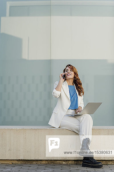 Businesswoman in white pant suit  sitting on bench  using laptop  talking on the phone