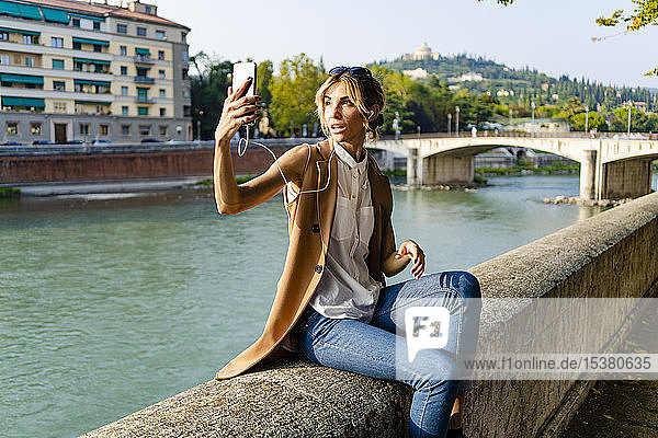 Frau mit Kopfhörern sitzt am Flussufer und macht ein Selfie