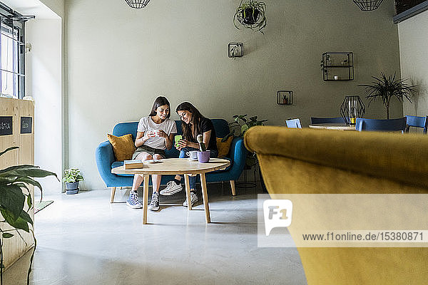 Zwei junge Freundinnen sitzen auf einer Couch in einem Café und teilen sich ein Handy