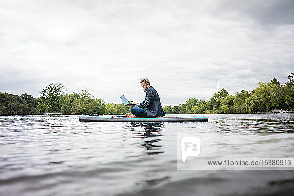 Geschäftsmann sitzt mit Laptop auf einem SUP-Brett auf einem See