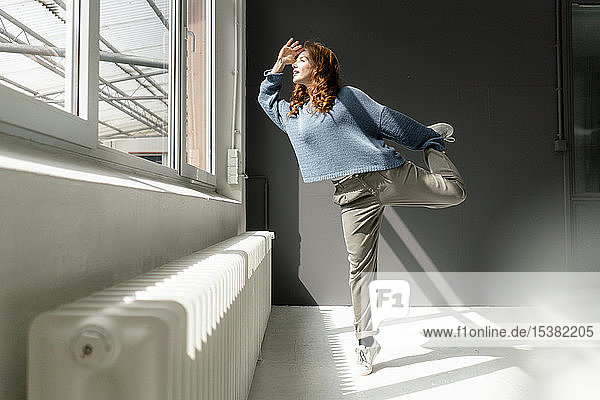 Redheaded woman standing on one leg in a loft looking out of window
