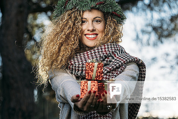 Portrait of smiling young woman with Christmas wreath on her head presenting Christmas present