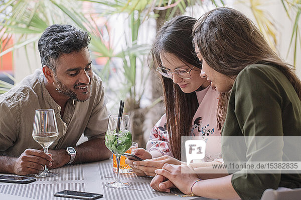 Freunde sitzen mit Getränken am Tisch und schauen auf das Smartphone