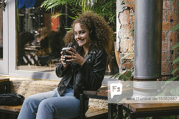 Portrait of smiling teenage girl sitting at outdoor cafe with coffee to go listening music with headphones