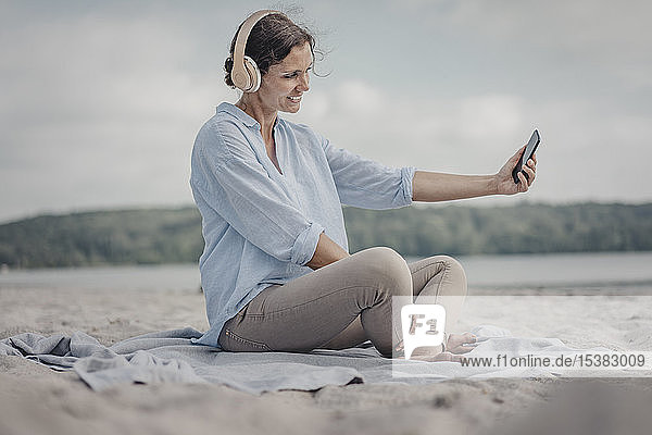 Frau sitzt am Strand  trägt Kopfhörer  nimmt Smartphone selbst