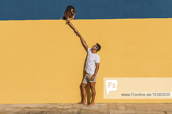 Junger Mann und Frau hinter einer gelben Wand halten sich an den Händen