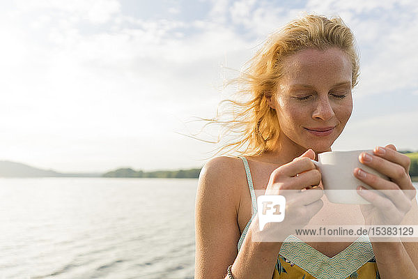 Junge Frau dmit geschlossenen Augen  die eine Tasse an einem See hält