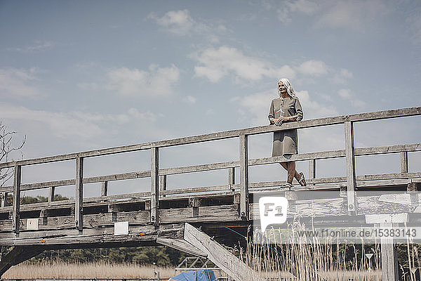 Ältere Frau steht auf Holzbrücke und hält Modell einer Windkraftanlage