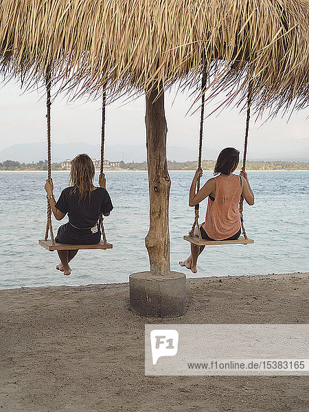 Rückenansicht zweier Frauen  die auf Schaukeln an der Strandpromenade sitzen  Gili Islands  Bali