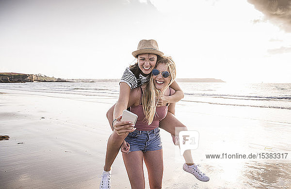 Two girlfriends having fun on the beach  carrying each other piggyback  taking smartphone selfies