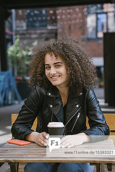 Portrait of smiling teenage girl sitting on bench with coffee to go and mobile phone