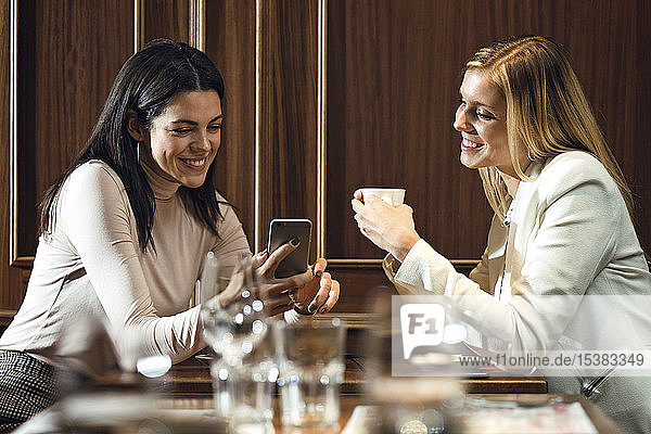 Zwei glückliche Frauen in einem Restaurant  die sich gemeinsam ein Smartphone anschauen