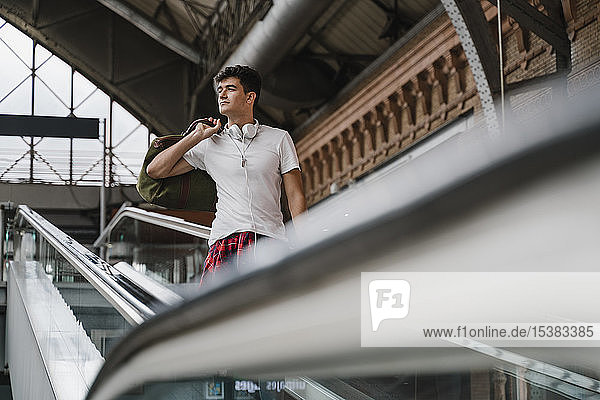 Junger Mann steht auf der Rolltreppe am Bahnhof und trägt Kopfhörer