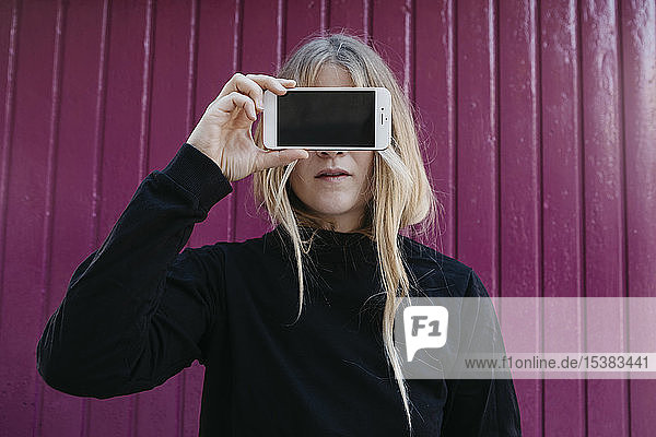 Porträt einer blonden jungen Frau  die mit einem Smartphone die Augen verdeckt