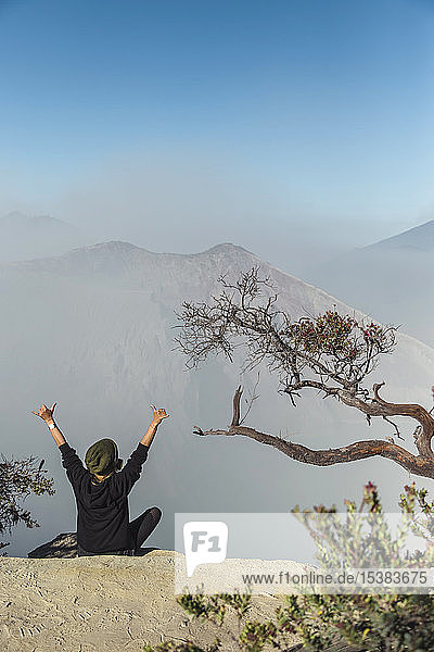 Frau mit Atemschutzmaske sitzt am Vulkan Ijen und hebt die Arme  Java  Indonesien