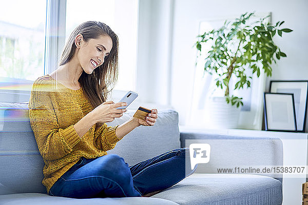 Schöne glückliche Frau bezahlt online mit Smartphone und Kreditkarte  während sie auf dem Sofa sitzt