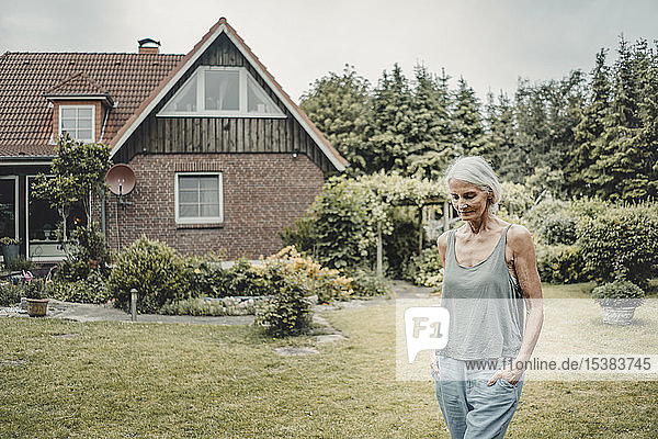 Ältere Frau geht in ihrem Garten spazieren  mit Händen in den Taschen