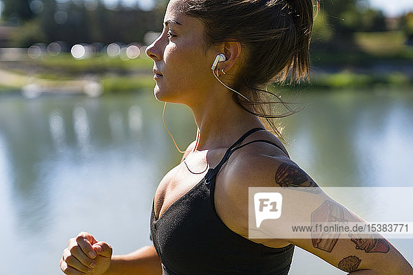 Tätowierte junge Frau mit Ohrstöpseln am Flussufer joggend