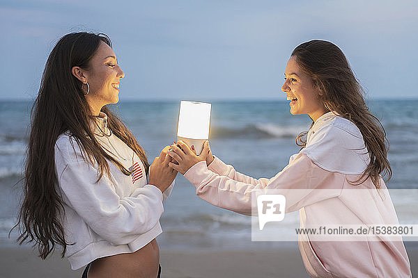 Zwei glückliche Freundinnen  die in der Abenddämmerung am Strand ein Führungslicht halten