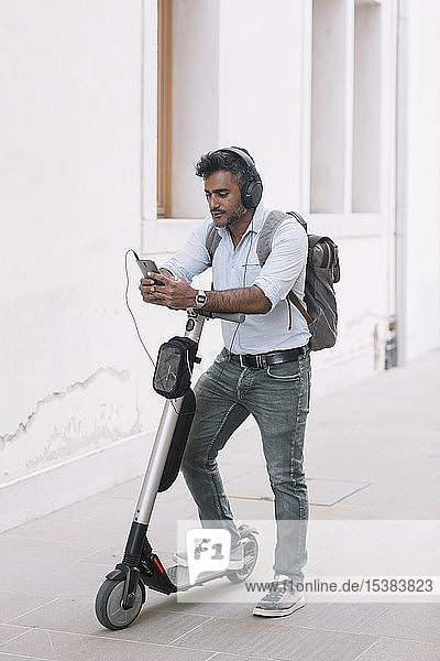 Casual businessman with headphones  smartphone and e-scooter in the city