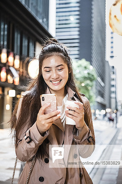 Glückliche junge Frau mit einem Getränk zum Mitnehmen und einem Smartphone in Ginza  Tokio  Japan