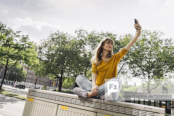 Junge Frau  die auf einer Kiste in der Stadt sitzt und ein Selfie