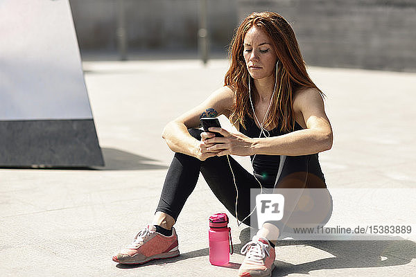 Sportliche junge Frau mit Kopfhörern macht eine Pause und benutzt das Smartphone im Freien
