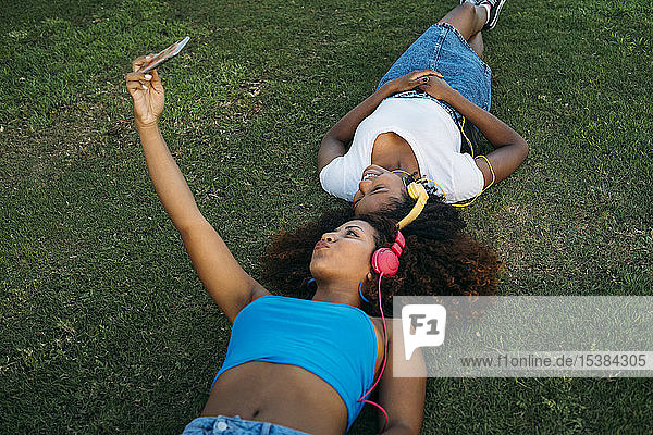 Zwei junge Frauen mit Kopfhörern liegen auf einer Wiese und nehmen sich mit dem Smartphone selbstständig