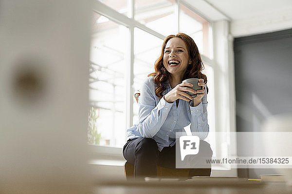 Bildnis einer lachenden rothaarigen Frau mit Teeschale auf der Rückenlehne sitzend auf einem Dachboden