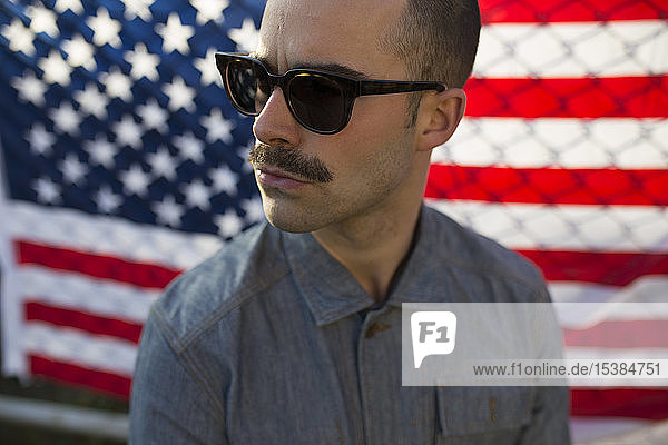 Junger Mann mit Sonnenbrille vor US-amerikanischer Flagge am Zaun
