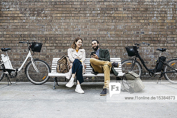Happy couple sitting on a bench next to e-bikes sharing cell phone