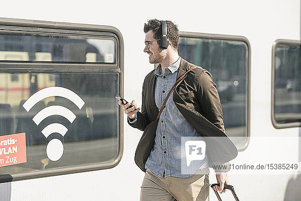 Lächelnder junger Mann mit Kopfhörern und Handy im Zug mit WLAN-Symbol