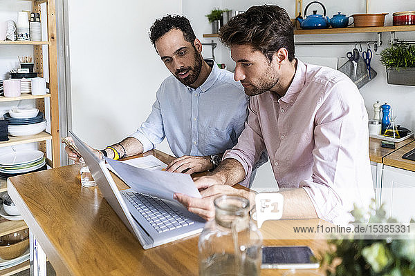 Zwei junge Männer arbeiten gemeinsam in der Küche  benutzen einen Laptop und diskutieren über Dokumente
