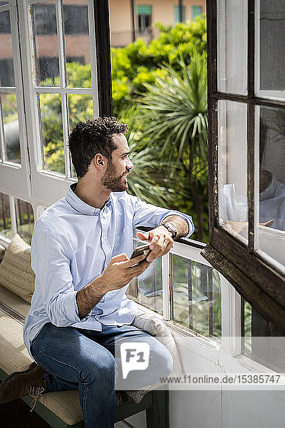 Junger Mann sitzt auf Bank am Fenster und benutzt Smartphone