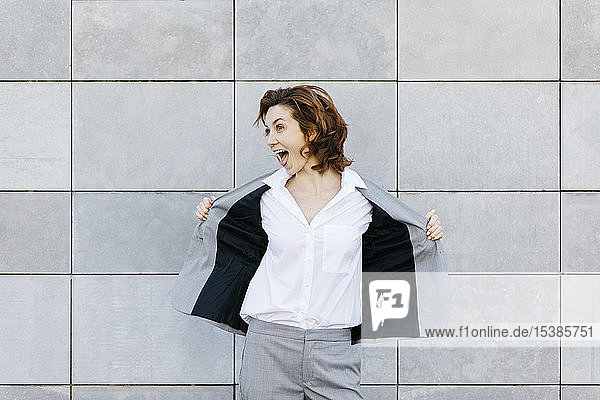 Porträt einer schreienden jungen Geschäftsfrau vor einer Wand mit grauen Kacheln  die ihre Jacke öffnet