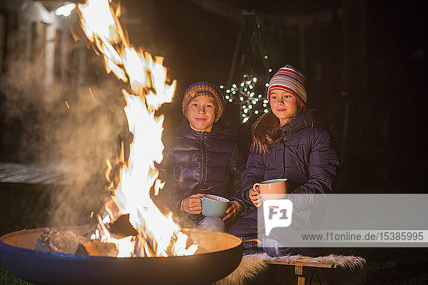 Junge und Mädchen mit Bechern sitzen nachts am Feuer