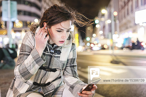 Spanien  Madrid  junge Frau in der Stadt  die nachts ihr Smartphone benutzt und Kopfhörer trägt