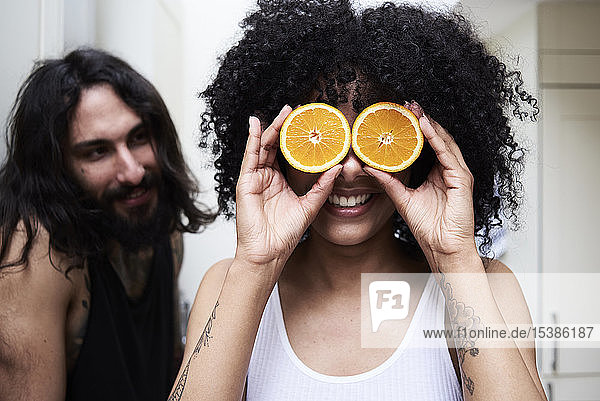 Happy young woman covering her eyes with oranges in kitchen