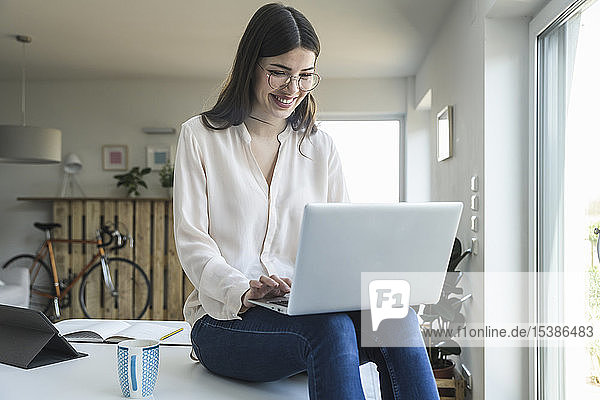 Lächelnde junge Frau sitzt zu Hause mit Laptop am Tisch