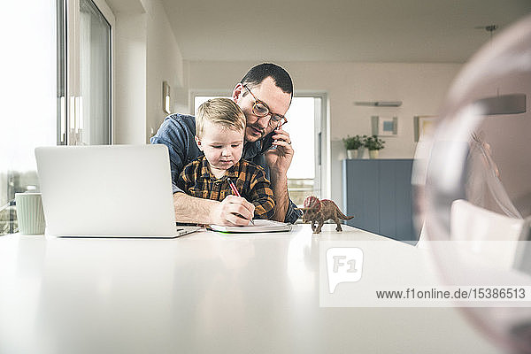 Busy father working at table in home office with son sitting on his lap